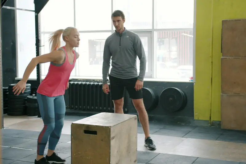 A woman jumping onto a wooden box in a gym while a coach watches