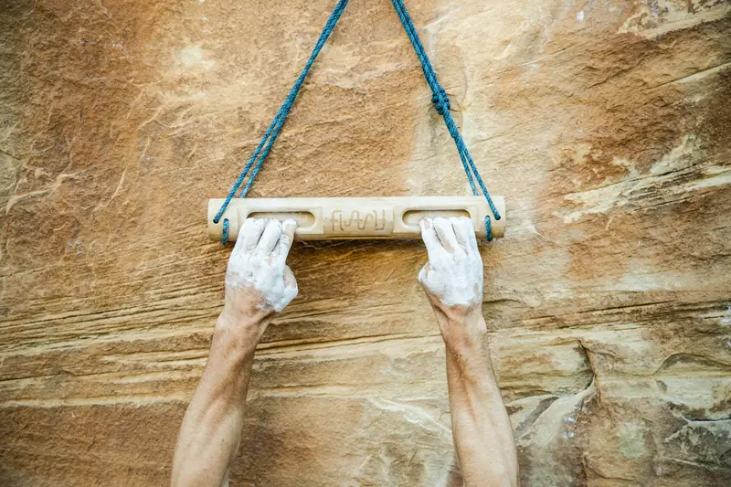 Chalked hands hanging from a wooden bar slung on ropes against sandstone