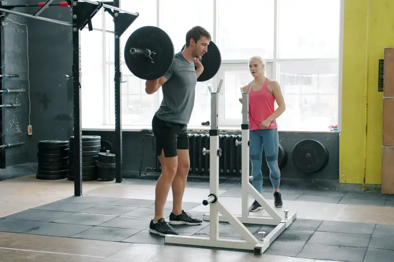 A man doing a barbell squat in a gym while a woman spots him