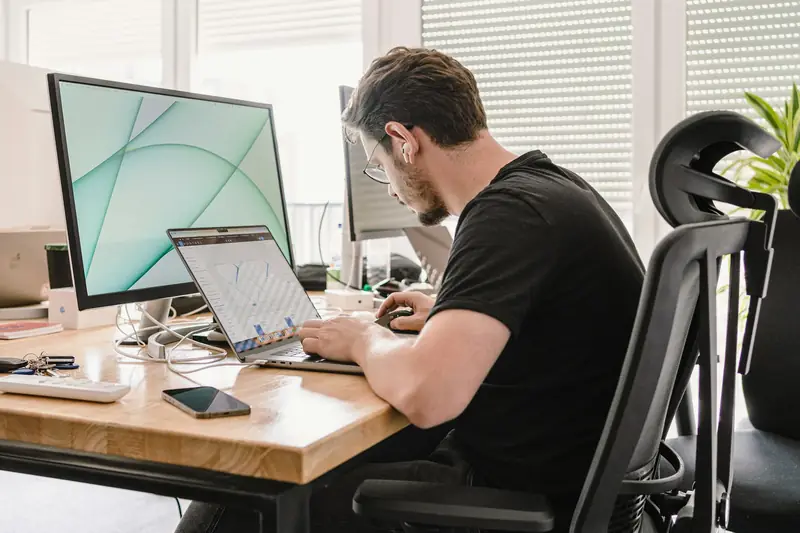 A man leaning in towards his laptop at a desk, a large monitor beside him