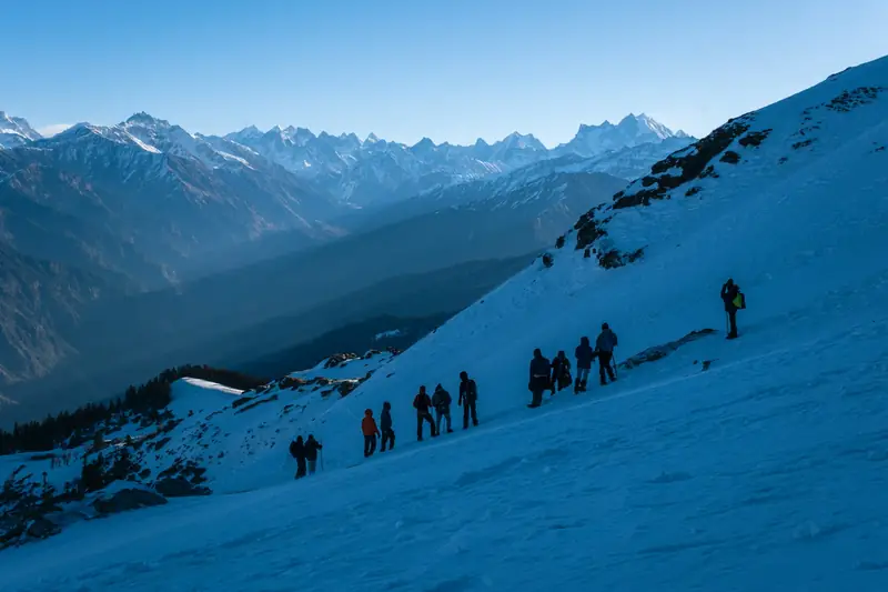A line of trekkers climbing a snowy slope with a high mountain range behind