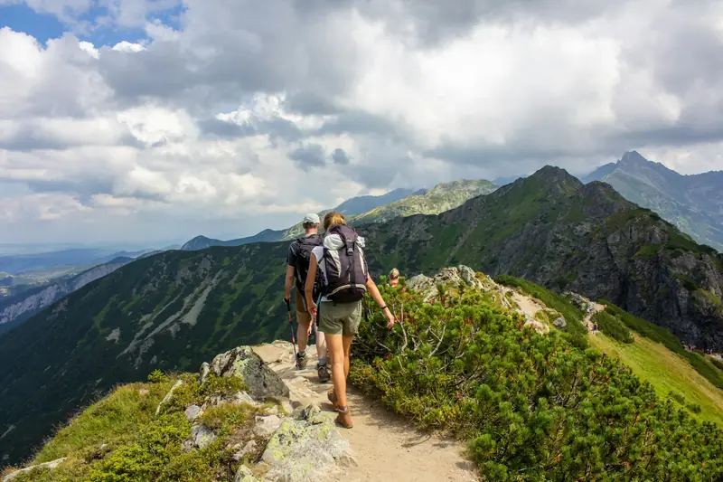 Hikers with backpacks walking up a narrow path along a green mountain ridge