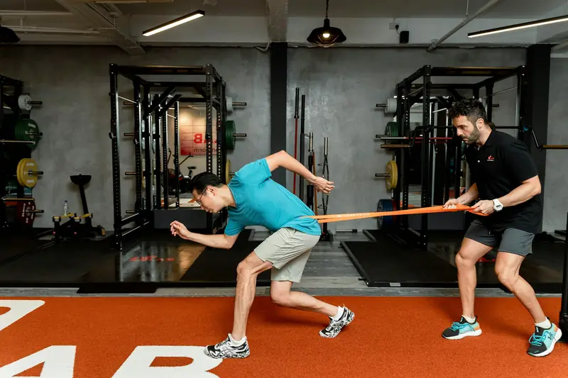 A trainer holding a resistance band while an athlete rotates against it in a gym