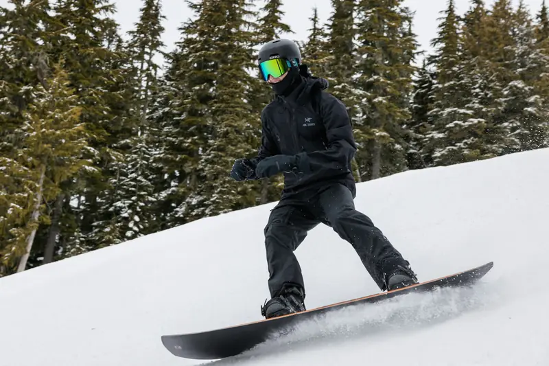 A snowboarder in black riding across a snowy slope lined with pine trees