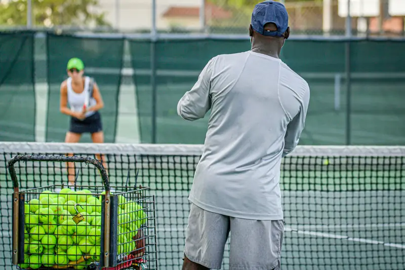 A coach feeding balls from a basket to a player waiting at the net
